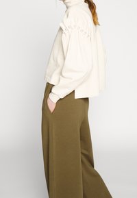 Krémový svetr s vysokým límcem, zdobený balónovými rukávy s perlovými detaily. V kombinaci s širokými olivově zelenými kalhotami.