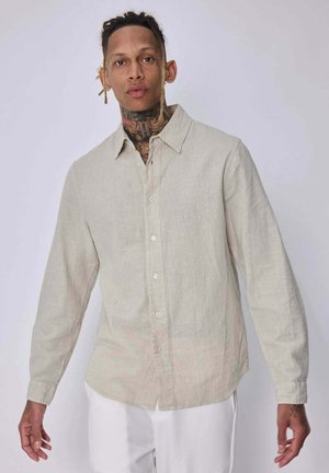 Homme avec des tatouages au cou portant une chemise beige à manches longues boutonnée et un pantalon blanc sur un fond clair uni.