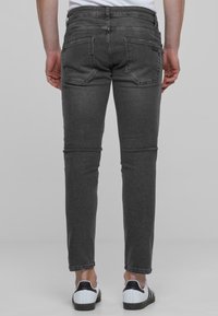 Pelēkie slim-fit džinsi ar piecām kabatām, izšūtu detaļu un tumšu mazgāšanu. Modelis valkā melnus un baltus sporta apavus, stāvot pret neitrālu fonu.