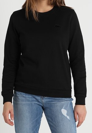 Kobieta ubrana w czarną, długorękawową bluzę z okrągłym kołnierzem, z małym logo krokodyla oraz jasnoniebieskie, podarte dżinsy.