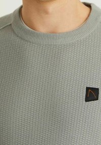 Grå texturerad tröja med ribbad halsringning, med en liten svart lapp med en orange triangel-logotyp på vänster bröstområde.