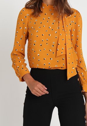 Vrouw draagt een mosterdgele blouse met zwart-witte polkadots, gecombineerd met hooggesnoerde zwarte broek met knoopdetails.