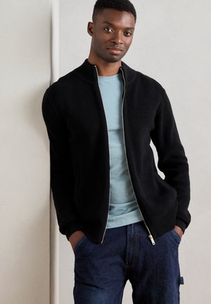 Svart ribbestrikket zip-up cardigan over en lys blå t-skjorte, kombinert med blå denimjeans. Enkel design med stående krage og kontrasterende teksturer.