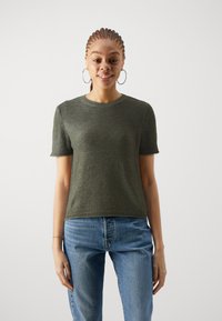 Dunkelgrüner, gestrickter Kurzarmpullover mit rundem Ausschnitt; kombiniert mit hellblauen Jeans. Schlichtes Design, strukturierter Stoff, keine Muster.