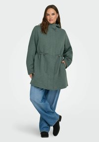 Zelena, kapuljača plašč z vrvico za prilagoditev v pasu in stranskimi žepi; narejen iz teksturirane tkanine. Usklajen z ohlapnimi modrimi kavbojkami in črnimi gležnjarji.