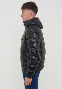 Giacca a vento nera con cappuccio, design trapuntato e zip gialla a contrasto. Toppa del logo sulla spalla sinistra. Texture liscia.