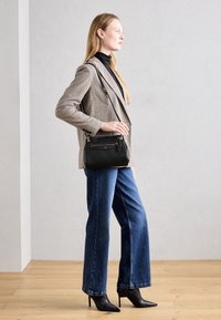 Donna che indossa un blazer a quadri, dolcevita nero, jeans blu e stivali neri con tacco, mentre tiene una piccola borsa a tracolla nera, in piedi di profilo.
