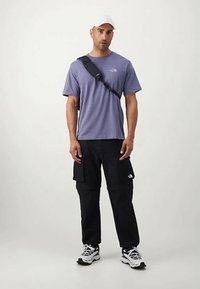 Mann iført en hvit caps, lilla kortermet skjorte, svarte cargo-bukser, svart crossover-veske og hvite joggesko, står mot en ensfarget bakgrunn.