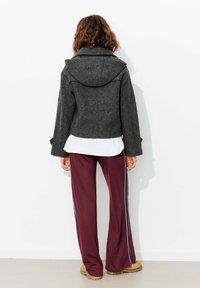 Veste en laine grise avec une capuche, manches larges et silhouette ajustée, associée à un pantalon bordeaux avec des bandes latérales, se tenant sur un fond neutre.