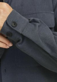 Mörkblå vävd tygskjortarm med två svarta knappar. Har ett texturerat mönster och en synlig ficka på övre delen.