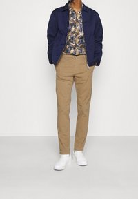 Man med beige byxor, vita sneakers, en blå jacka över en mönstrad skjorta med knappar, står mot en ljus enfärgad bakgrund.