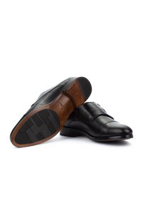 Par de zapatos de vestir de cuero negro con hebillas, uno mostrando la suela lisa con agarre de goma y detalles en el tacón sobre fondo blanco.