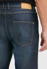 Vaqueros de denim azul oscuro con un parche de cuero en la cintura, costuras visibles y dos bolsillos traseros con costuras en contraste.