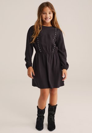 Ein junges Mädchen lächelt, trägt ein schwarzes Langarmkleid mit silbernen Nieten und schwarze, mit Nieten besetzte Stiefel bis zur Mitte der Wade, und steht vor einem einfarbigen Hintergrund.