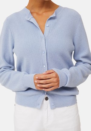 Persona che indossa un cardigan azzurro chiaro a coste con bottoni e maniche lunghe, e pantaloni bianchi, con le mani giunte in vita.