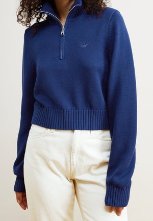 Mujer con un jersey de punto azul marino con cuello de media cremallera y puños acanalados, combinado con pantalones de cintura alta color blanco roto.