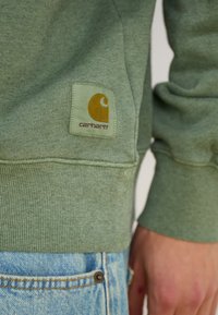 Green sweatshirt with ribbed cuffs and hem, featuring a square logo patch in yellow and orange, worn over light blue denim.