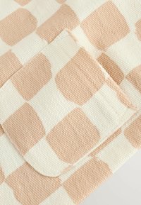 Beige en perzikkleurig geruit stof met een gestructureerde oppervlakte. Heeft een zakflap die het patroon en het materiaal benadrukt.
