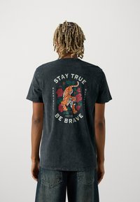 Černé bavlněné tričko s grafikou tygra a růží na zádech, s textem "ZŮSTAŇTE PRAVDIVÍ A ODVAŽNÍ" v bílé barvě.