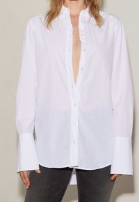 Chemise blanche à boutons avec des manches longues et un décolleté en V profond, dotée d'une patte froncée et de larges poignets, fabriquée dans un tissu léger.