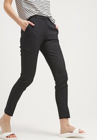 Personne portant un pantalon noir slim, une chemise rayée et des sandales blanches à enfiler, debout les mains dans les poches devant un fond uni.