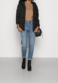 Sort pufferjakke med hætte, brun t-shirt, lyseblå jeans og sorte hælede støvler, der viser et afslappet lag-på-lag outfit.