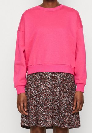 Persoon die een felroze sweatshirt met lange mouwen en een donker bloemenpatroonsrok draagt, staat tegen een effen achtergrond.