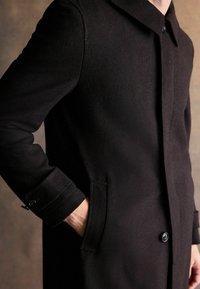 Osoba w czarnym zapinanym płaszczu, z jedną ręką w kieszeni, na neutralnym tle.