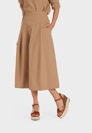 Khaki-kleurige midi-rok met een aansluitende taille en wijde pijpen, met zichtbare stikdetails. Gepaard met bruine plateausandalen.