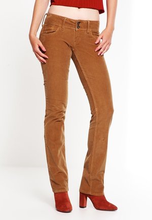 Frau trägt schmal geschnittene braune Cordhosen und rote Stiefeletten mit Absatz, steht mit entspannten Händen an den Seiten.