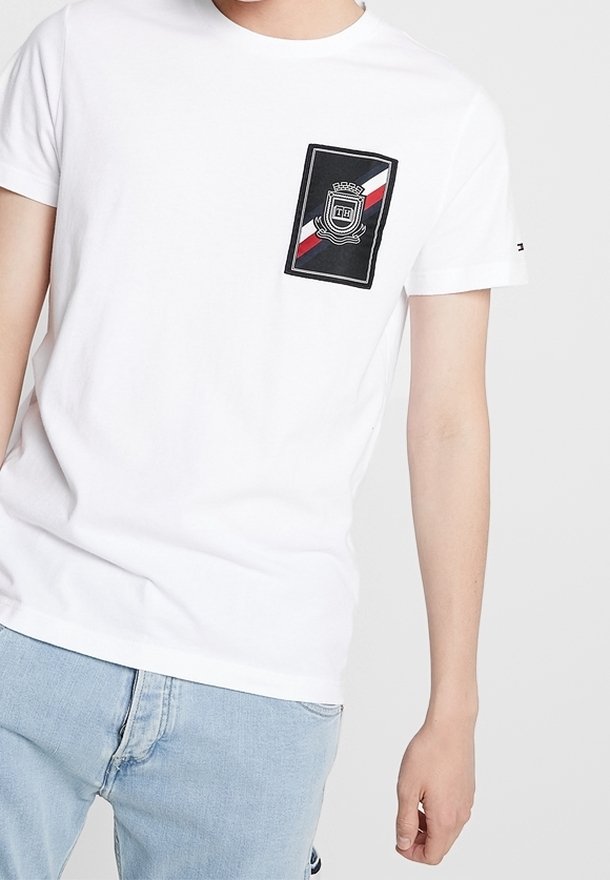 Camiseta de algodón blanca con un bolsillo negro con un diseño de escudo y bandas diagonales rojas y azules. Manga corta y cuello redondo.