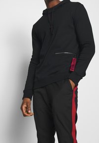 Hoodie noir avec une poche zippée à l'avant et une étiquette logo rouge. Tissu lisse, capuche avec cordon de serrage et coupe décontractée. Associé à un pantalon noir avec des rayures rouges.