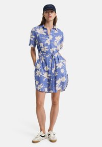 Blå blomsterkjole med knapper, kortermet, midjebelte og sidelommer. Brukt med hvite joggesko med svarte detaljer.
