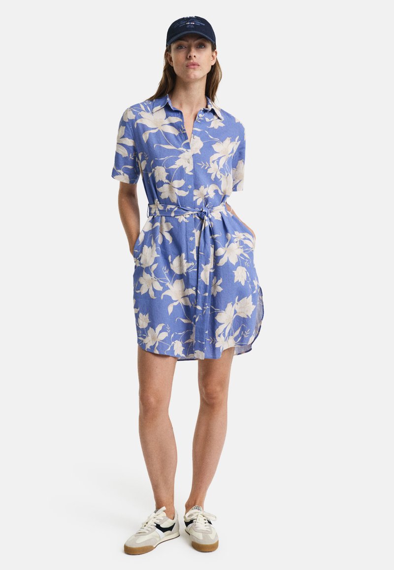 Blå blomsterkjole med knapper, kortermet, midjebelte og sidelommer. Brukt med hvite joggesko med svarte detaljer.