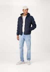 Giacca a vento navy con motivo a chevron, cerniera frontale e cappuccio. Indossata con una t-shirt bianca e jeans azzurri sbiaditi. Cappellino beige.