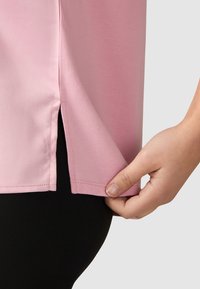 Tee-shirt rose à manches courtes en tissu lisse, avec des fentes sur les côtés à l'ourlet. Une main tient le bas, révélant un matériau noir en dessous.