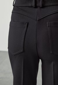 Black tailored trousers featuring a smooth texture, traditional back pockets, and a fitted design with sharp stitching details.