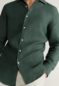 Camisa de lino verde oscuro con cierre de botones en la parte delantera, mangas largas y cuello clásico. Cuenta con botones blancos y una textura ligera.
