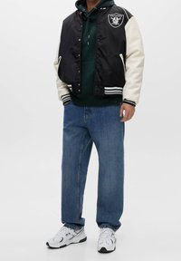 Stijlvolle zwarte en crème bomberjack met strepen, met een logo, gedragen over een groene hoodie, gecombineerd met losse blauwe jeans en witte sneakers.