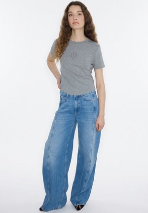 Jeune femme aux longs cheveux bouclés portant un t-shirt gris étincelant et un jean large bleu, debout avec une main sur la hanche.