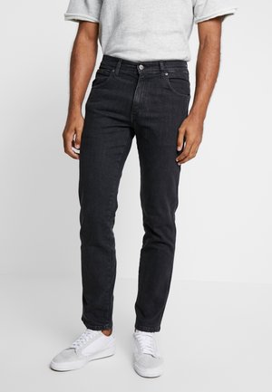 Homme portant un jean slim noir, des baskets blanches basses et un t-shirt gris clair à manches retroussées, debout sur un fond blanc uni.
