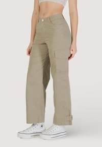 Pantalones cargo beige de corte relajado, con grandes bolsillos laterales, cierre de botón y dobladillos ajustados. Combinados con zapatillas blancas.
