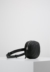 Sac rond en cuir noir avec une texture matelassée, une fermeture éclair argentée et une bandoulière réglable, posé sur une surface blanche.