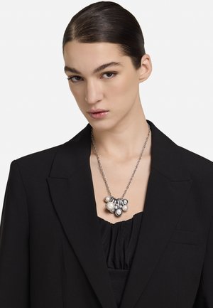 Collana a catena tono argento con un grappolo di perle bianche e accenti rotondi in argento, indossata su una maglia nera e un blazer.