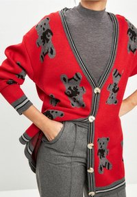 Cardigan rosso con motivo di orsetti grigi, design a V, dettagli a strisce grigie e bottoni. Indossato sopra un maglione a collo alto grigio.