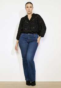 Črna cvetna prosojna bluza z V-izrezom, v kombinaciji z visokimi temno modrimi širokimi hlačami. Model nosi črne škornje z visokim petam. Enolična podlaga.