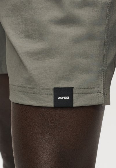 Κοντινή λήψη του τελειώματος μιας χακί σορτς με μαύρη ετικέτα της μάρκας ASPESI σε σκουρόχρωμο καφέ δέρμα.