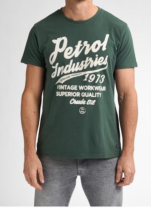 Mann trägt ein dunkelgrünes T-Shirt mit weißem Text "Petrol Industries 1973 Vintage Workwear Superior Quality Crude Oil" und graue Jeans.