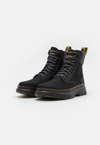 Paire de bottines noires à lacets avec semelles épaisses, surpiqûres jaunes et languettes de tirage brandées à l'arrière sur fond blanc.