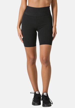 Woman standing wearing black high-waist biker shorts and black sneakers with laces against a white background.